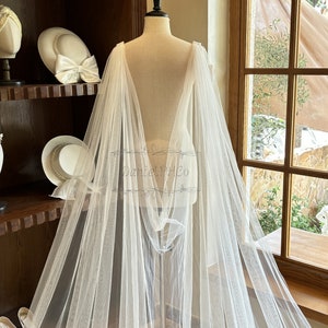 May include: A white tulle bridal cape with a long train, draped over a dress form. The cape is sheer and delicate, with a flowing design. The cape is perfect for adding a touch of elegance to any wedding dress.