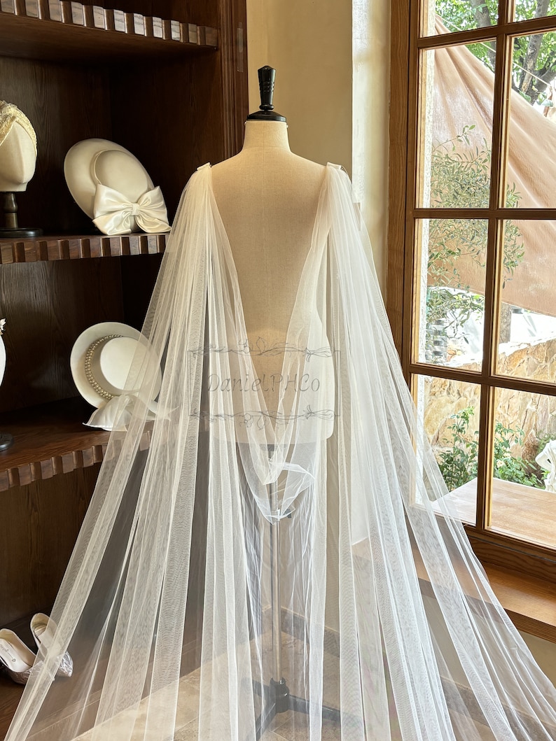 May include: A white tulle bridal cape with a long train, draped over a dress form. The cape is sheer and delicate, with a flowing design. The cape is perfect for adding a touch of elegance to any wedding dress.