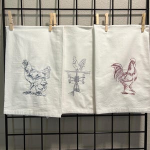 May include: Three white tea towels with embroidered designs. The first towel features a hen and two chicks, the second towel features a rooster weather vane, and the third towel features a rooster.