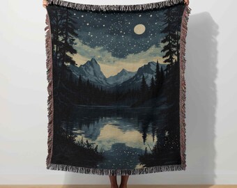 May include: A woven throw blanket featuring a serene landscape of a lake, mountains, and a starry night sky. The design includes a full moon, pine trees, and a reflection in the water. The blanket has a fringed edge.