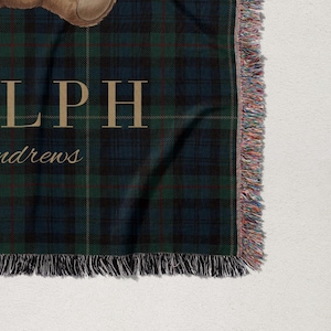 a close-up view of a plaid blanket with fringed edges, and a small image of a teddy bear on it.