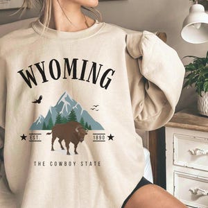 Könnte beinhalten: Ein beiger Pullover mit einem Grafikdesign mit dem Text "WYOMING", einer Bergkette, einem Bison und dem Text "The Cowboy State".