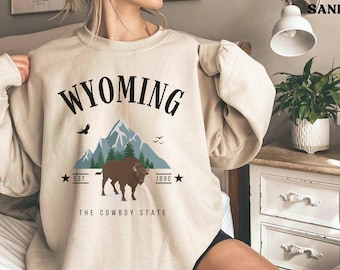May include: A beige sweatshirt with a graphic design featuring the text "WYOMING", a mountain range, a bison, and the text "The Cowboy State".