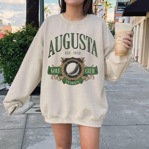 May include: A cream-colored sweatshirt with a green and white graphic print that says "Augusta Est. 1932 Golf Club Georgia".