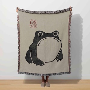 May include: A beige throw blanket featuring a large black frog illustration. The frog has a minimalist design with large eyes and a wide mouth. The blanket has a fringed edge and a red stamp-like design in the upper left corner.
