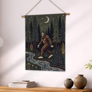 May include: A tapestry wall hanging featuring a Bigfoot illustration walking near a river in a forest. The artwork includes a crescent moon and stars in the night sky, with a wooden rod and string for hanging. The color palette includes shades of green, brown, and blue.