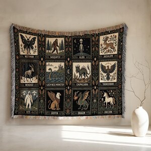 May include: Woven tapestry blanket featuring a grid of mythical creatures including Mothman, Sasquatch, and a Unicorn. The design incorporates a black background with floral accents and fringe edges. Each creature is labeled with its name.