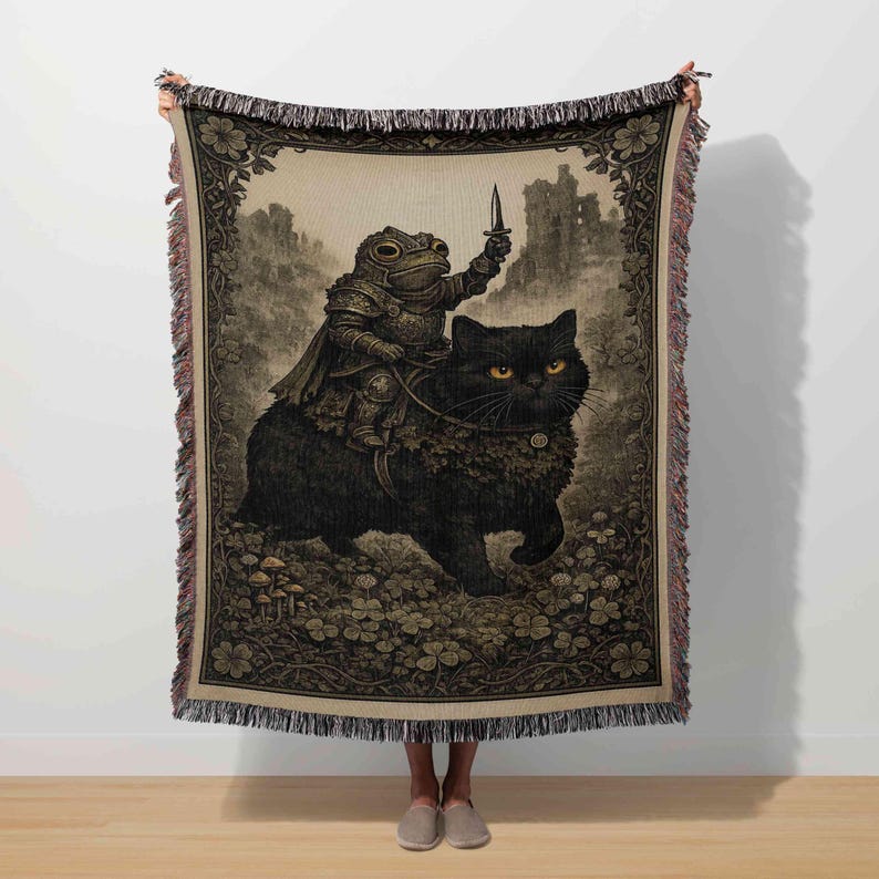 A person is holding a large tapestry or blanket featuring a detailed illustration of a frog riding a black cat, with the frog holding a sword.