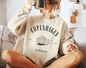 May include: A beige sweatshirt with a black graphic print of a building and the text "Copenhagen Est. 1167 Denmark".