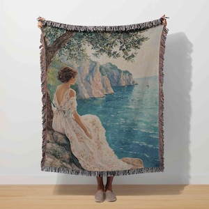 May include: Woven throw blanket featuring a scenic painting of a woman in a white dress, seated on a rock overlooking the sea. The artwork includes a tree, cliffs, and a sailboat, with a fringed border.