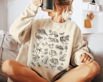 May include: A cream-colored sweatshirt with a black and white illustration of various woodland creatures and plants. The illustration includes a deer, a squirrel, a bird, a mushroom, a dragonfly, and a snail.