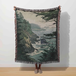May include: A woven throw blanket featuring a coastal landscape scene. The design depicts a rocky shoreline with evergreen trees, a flowing river, and a misty sky. The blanket has a fringed edge and is primarily green, gray, and blue.