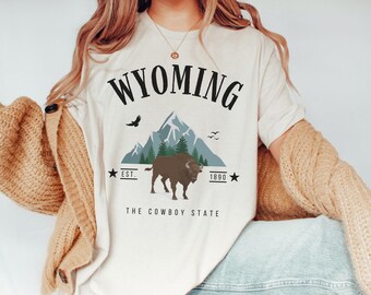 May include: A white t-shirt with a black graphic design that says "WYOMING" with a mountain range, a bison, and the text "EST. 1890 THE COWBOY STATE".