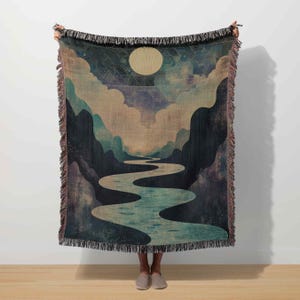 May include: A woven throw blanket featuring a landscape design with a river winding through mountains under a full moon. The color palette includes blues, greens, and beige, with fringed edges.