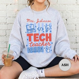 May include: A light gray crewneck sweatshirt featuring the red text Mrs. Johnson and Tech teacher, accompanied by blue graphic icons of a laptop, a lightbulb, a computer keyboard, and a mouse, all set against a background of vertical blue stripes.