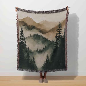 May include: A woven throw blanket featuring a watercolor landscape of a forest and mountains in shades of green, brown, and beige. The design depicts a misty forest scene with tall trees and distant mountain ranges. The blanket has a fringed edge.