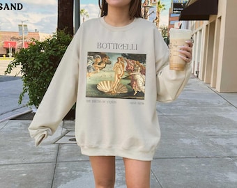 May include: A cream-colored sweatshirt with a graphic print of Sandro Botticelli's "The Birth of Venus" painting. The text "Botticelli" and "The Birth of Venus" is printed below the image. The text "1482-1486" is printed below the text "The Birth of Venus".