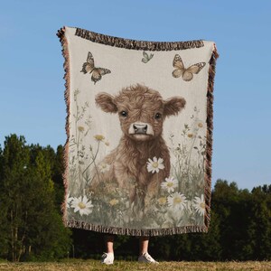 A person is standing in a grassy field, holding a blanket that features a cow and butterflies.