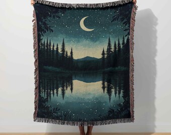 May include: A woven throw blanket featuring a nighttime forest scene. The design includes a crescent moon, a starry sky, and a lake reflecting the trees. The blanket has a fringed edge and is primarily dark blue and green.