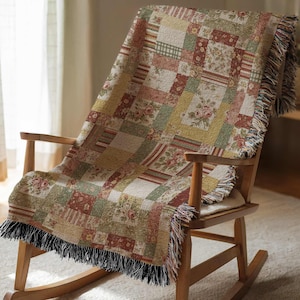 May include: A patchwork throw blanket draped over a wooden rocking chair. The blanket features a mix of floral and geometric patterns in shades of green, red, and cream. The edges are finished with a black fringe.