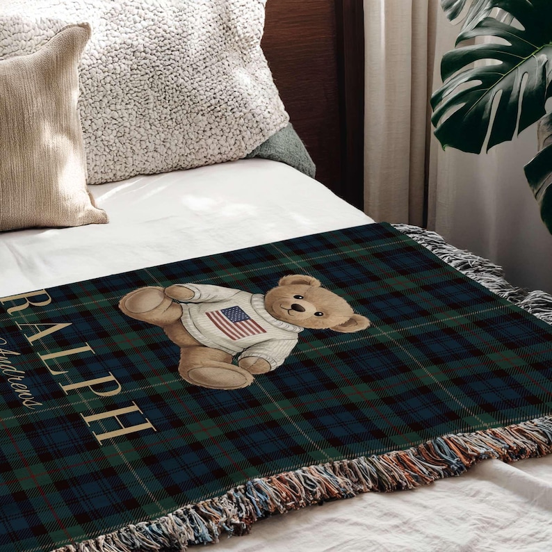 a cozy bedroom scene with a bed covered in a plaid blanket that has a teddy bear wearing a sweater with an American flag design. The bed is surrounded by various decorative pillows, and there is a potted plant in the background.