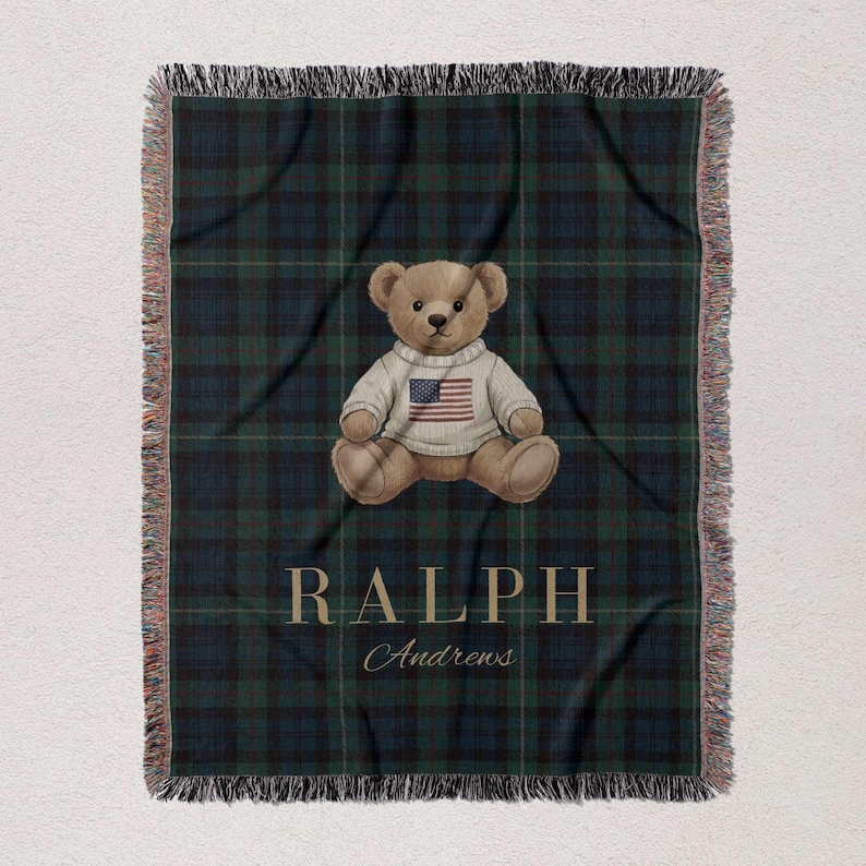 A teddy bear wearing a sweater with an American flag on it, sitting on a plaid blanket with the name "RALPH Andrews" printed on it.