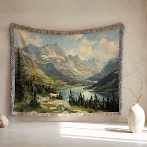 May include: A woven tapestry depicting a mountain landscape with a lake and a bighorn sheep. The tapestry has a fringed edge and is displayed on a wall. The colour palette includes blues, greens, and earth tones. The tapestry measures approximately 150 cm x 200 cm.