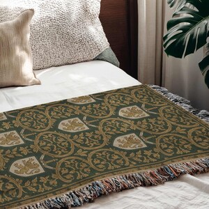 May include: A green throw blanket with a repeating pattern of gold heraldic crests and foliage. The blanket has a fringed edge with multiple colors and is displayed on a bed with white sheets and pillows.