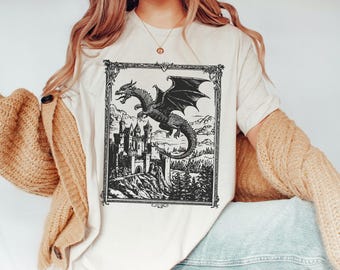 May include: A white t-shirt with a black and white illustration of a dragon flying over a castle. The illustration is framed in a black frame with a decorative border.