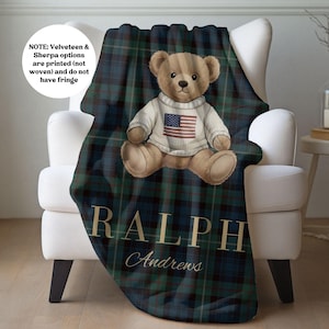 May include: A dark green and navy blue plaid blanket featuring a central illustration of a brown teddy bear wearing a white sweater with an American flag graphic. The text RALPH Andrews is printed in gold lettering below the bear. NOTE: Velveteen & Sherpa options are printed (not woven) and do not have fringe.