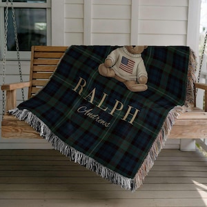 A plush teddy bear wearing a sweater with an American flag design sits on a wooden bench, with a plaid blanket featuring the word "RAPHP" and the word "Chapman" printed on it.