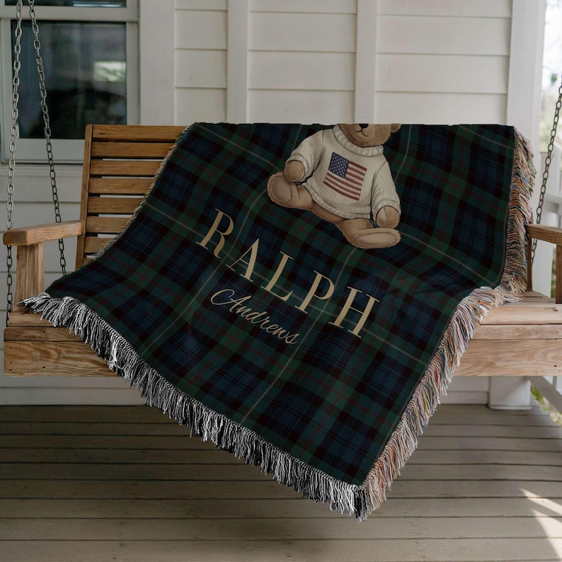 A plush teddy bear wearing a sweater with an American flag design sits on a wooden bench, with a plaid blanket featuring the word "RAPHP" and the word "Chapman" printed on it.