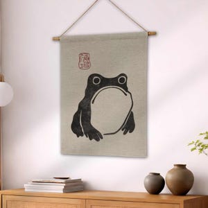 May include: A beige wall hanging featuring a black frog illustration. The frog has large eyes and a wide mouth. A red stamp-like symbol is in the upper left corner. The wall hanging is suspended from a wooden rod with a natural fiber cord.