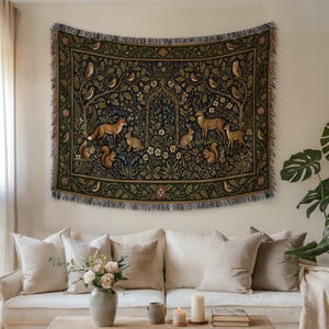 May include: A decorative tapestry depicting a woodland scene with foxes, deer, rabbits, squirrels, and owls. The tapestry has a dark green backdrop with intricate floral and tree patterns, and a fringed border. It is displayed on a wall above a white sofa.