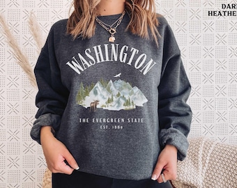 May include: Dark heather grey sweatshirt with a white graphic print of a mountain range and a moose. The text "WASHINGTON THE EVERGREEN STATE EST. 1889" is printed below the graphic.