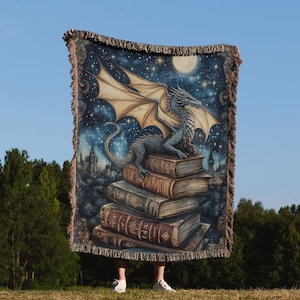 May include: A woven throw blanket featuring a fantasy scene. A blue dragon with tan wings sits atop a stack of antique books under a starry night sky with a full moon. The blanket has a fringed edge and a detailed illustration.