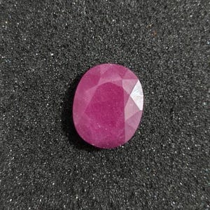 May include: A faceted, oval-shaped ruby gemstone. The gem is a deep pinkish-red color and has multiple facets that reflect light. The gemstone is set against a dark, textured background.
