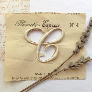 以下が含まれることがあります： Plumetis Express No 4およびMade in Franceの文字が記されたヴィンテージ風のベージュ色の紙ラベルの中央に、クリーム色の刺繍された文字Cのアップリケが配置されています。特許の詳細が記載された紙の上に、乾燥したラベンダーの小枝が斜めに置かれています。