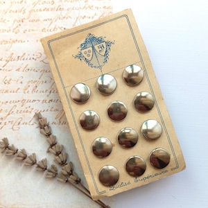 Può includere: Un set di undici bottoni rotondi in metallo color oro disposti su un cartoncino rettangolare color beige. Il cartoncino presenta un logo con stemma blu in alto e la scritta Qualité Supérieure in basso, accompagnato da un rametto di lavanda.