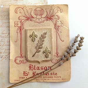 Può includere: Una carta rettangolare con uno stemma stampato in rosso con la parola CANNES, sormontato da uno scudo con due gigli dorati e una spiga di grano. Il testo Blason Hte Fantaisie è stampato sotto, accompagnato da steli di lavanda essiccata.