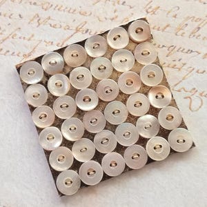 May include: A collection of round, iridescent mother-of-pearl buttons with two holes, arranged on a square of textured, brown paper. The buttons have a subtle sheen and are evenly spaced, creating a neat pattern. The background has faded script.
