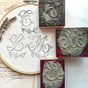 May include: Three rectangular wooden rubber stamps featuring ornate, engraved cursive letters E, M, and L, each decorated with floral motifs. Beside them, an embroidery hoop holds white fabric printed with the same three letter designs.