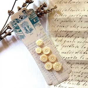 Puede incluir: Un marcapáginas de estilo vintage con un diseño azul y crema, con una imagen religiosa y texto en francés. Varios botones crema con forma de flor están adjuntos. Ramas de lavanda secas y escritura antigua en el fondo.