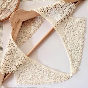 Puede incluir: Cuello de encaje color crema con un delicado diseño floral, exhibido en una percha de madera. El cuello tiene un borde festoneado y probablemente sea vintage o hecho a mano. El intrincado diseño sugiere un enfoque en el detalle y la artesanía.