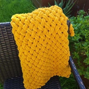 Peut inclure: Une couverture jaune moutarde tricotée à la main avec un motif de tissage épais. La couverture est drapée sur une chaise en osier noire, avec un feuillage vert en arrière-plan. La texture de la couverture est douce et accueillante.