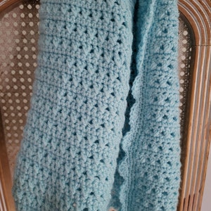 Peut inclure: Une couverture en crochet bleu clair avec un bord festonné. La couverture est accrochée à une chaise en bois.