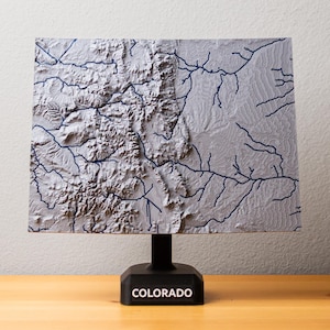 Könnte beinhalten: Eine graue 3D-Reliefkarte von Colorado mit detaillierter Topographie und blauen Flusslinien. Die Karte ist auf einem schwarzen Sockel mit dem Staatsnamen in Weiß montiert. Das Produkt wird auf einer Holzoberfläche präsentiert.