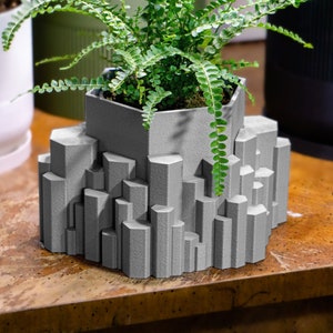 May include: A gray geometric planter with a fern plant growing inside. The planter is made of a textured material and has a unique, modern design.