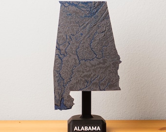 Può includere: Una mappa in rilievo grigia e blu dell'Alabama, montata su un supporto nero. Il supporto ha il nome "ALABAMA" in testo bianco. La mappa è dettagliata, mostrando la topografia dello stato.
