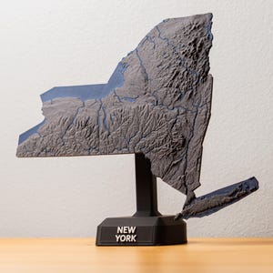 Pode incluir: Um mapa topográfico impresso em 3D do estado de Nova Iorque, em tons de cinza e azul, montado em um suporte preto. A base do suporte diz "NEW YORK" em texto branco. O mapa mostra o terreno do estado.